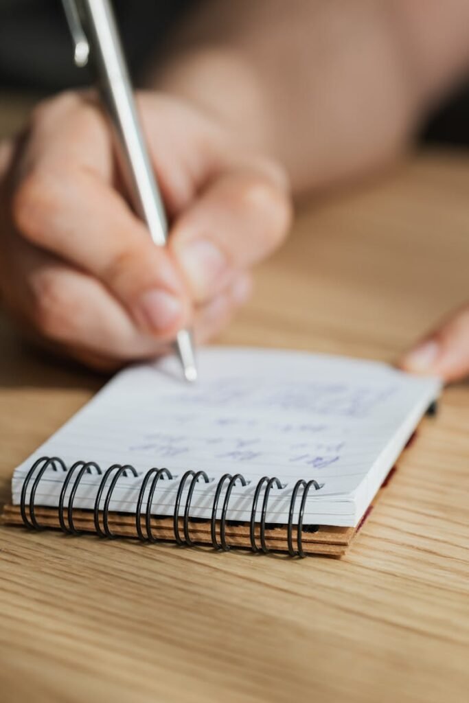 From above of crop faceless male student writing with pen in notebook at wooden desk during lesson